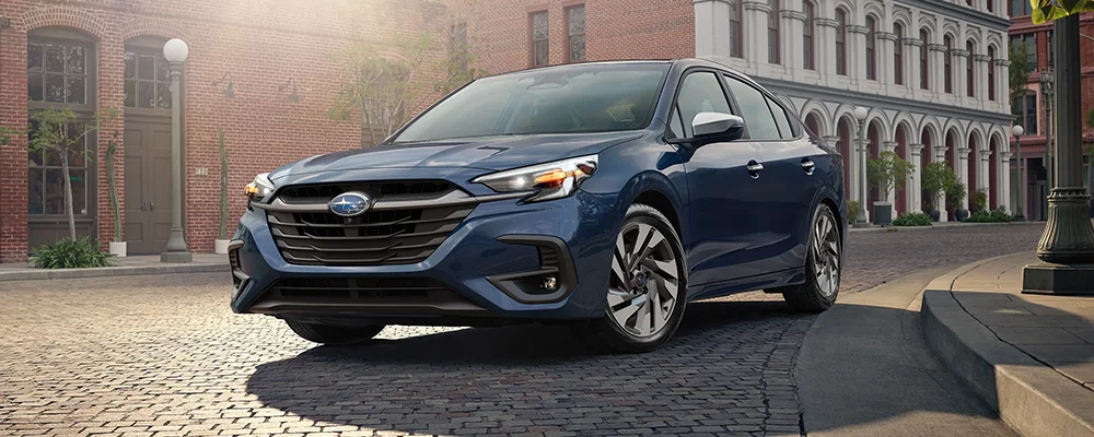 A blue Subaru Legacy AWD car parked on a cobblestone street in front of a brick building.